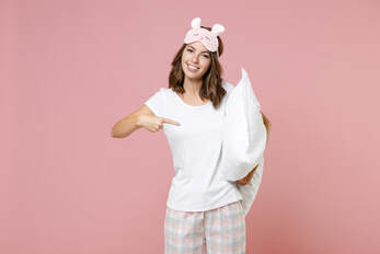 Woman in pajamas with an blindfold on her head holing a pillow under one hand and pointing at it with the other